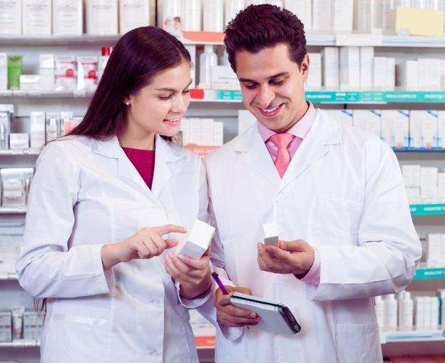 Pharmacists in lab coats discussing products in a pharmacy, holding medication boxes.