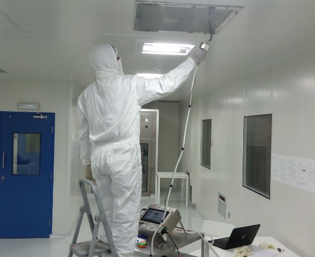 Technician inspecting ceiling ventilation system in a cleanroom with a sampling device.